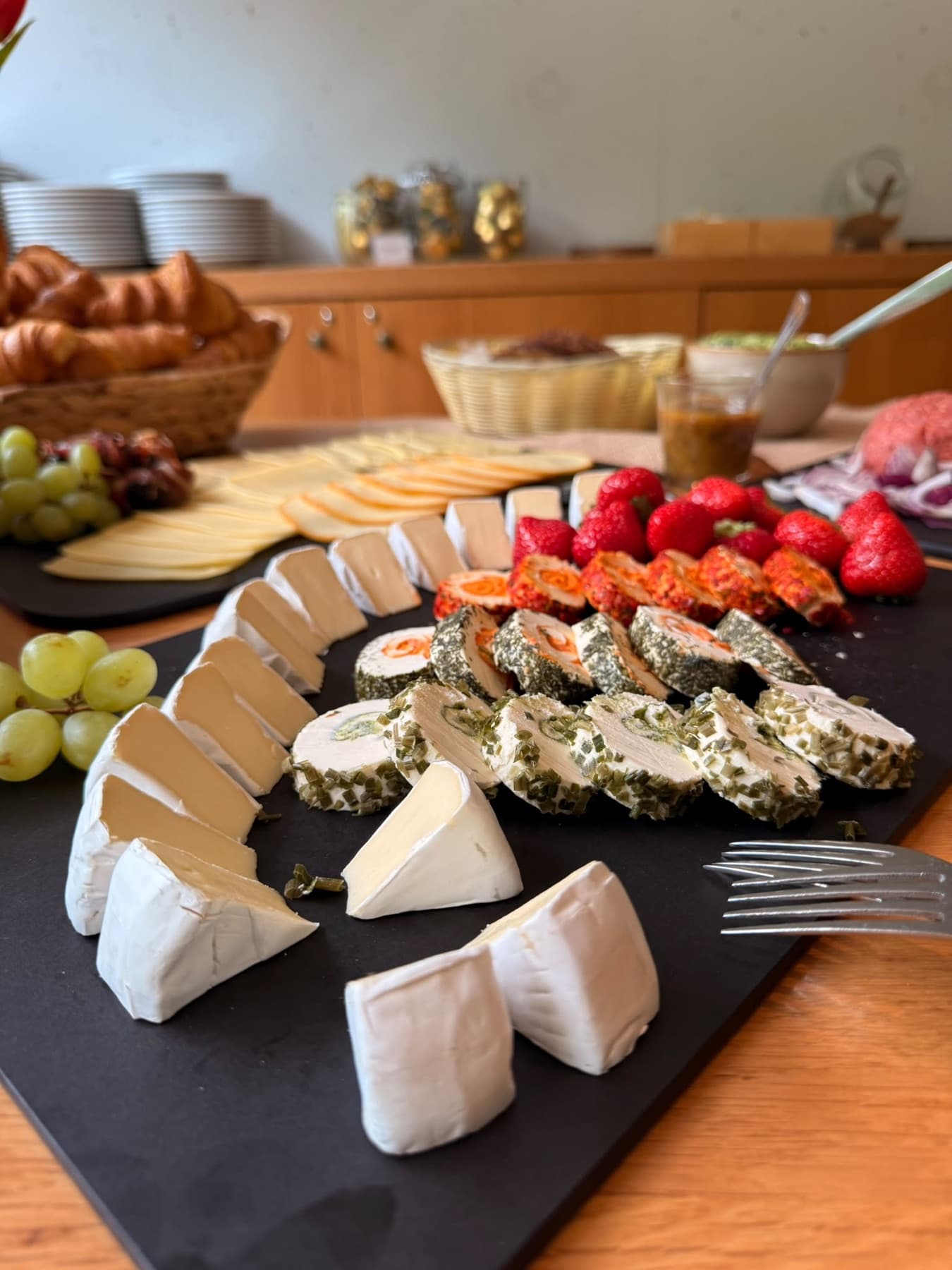 Käsevariation mit Weintrauben und Beeren, dahinter ein Korb mit frischem Gebäck