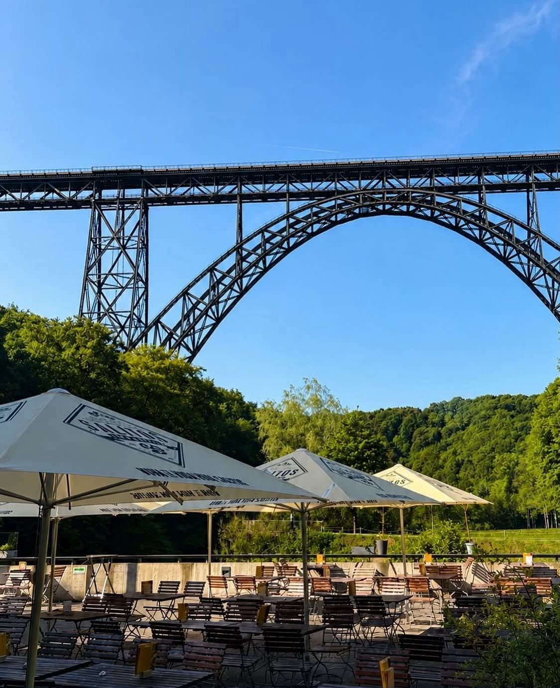 Terrasse des Hauses Müngsten mit Holztischen und hellen Sonnenschirmen, darüber der Bogen der Müngstener Brücke vor blauem Himmel
