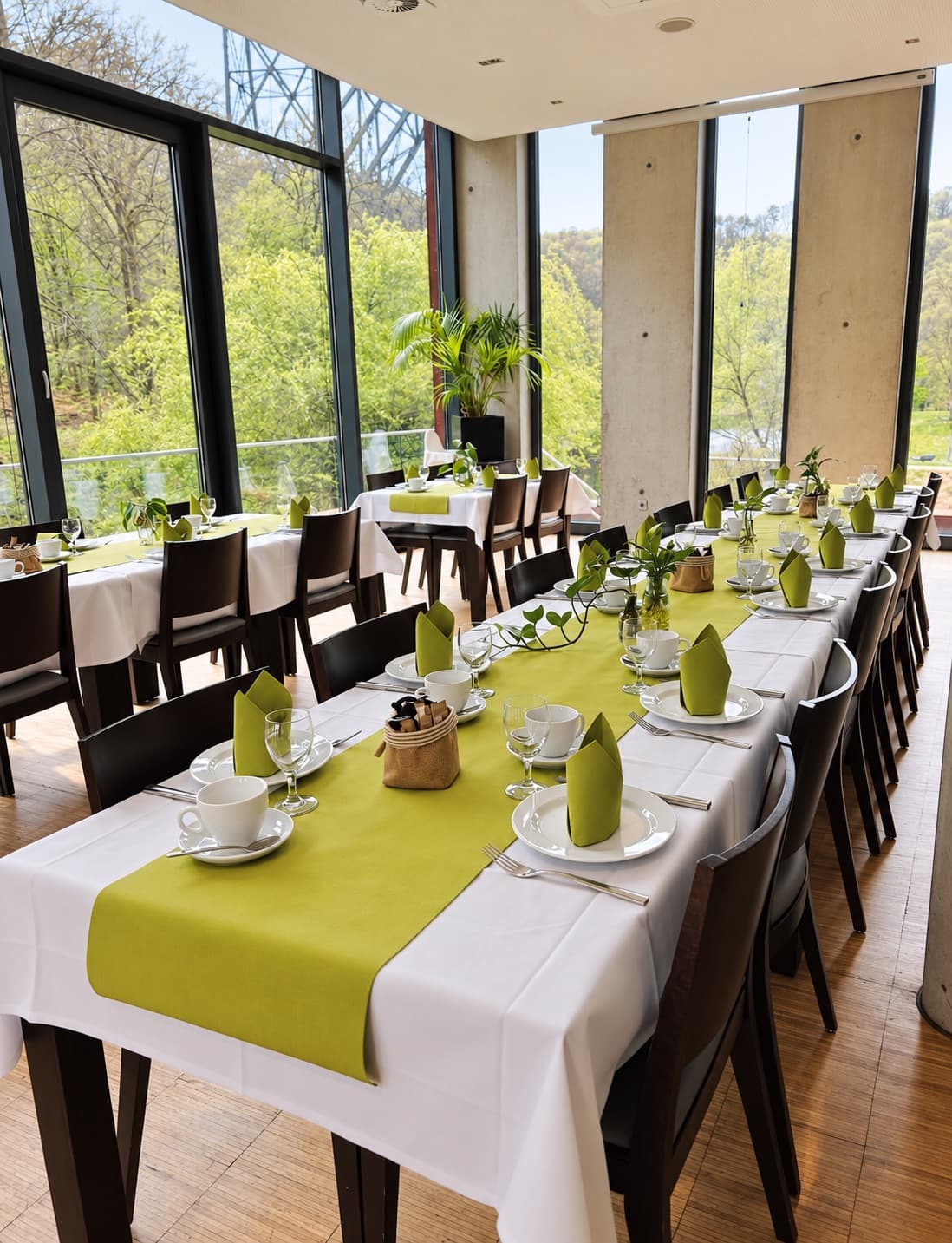 Lange Tafel im Raum Naturblick, weiß eingedeckt mit grünen Läufern und Servietten, dahinter die raumhohe Fensterfront zum bewaldeten Tal